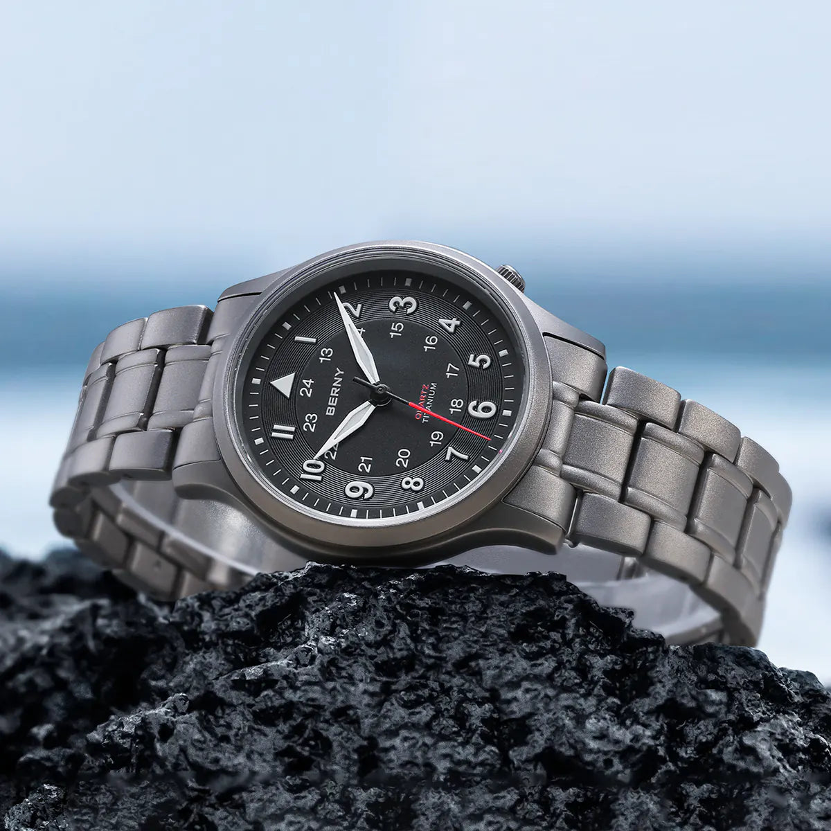 This Berny watch is elegantly displayed on a rocky surface with a background of the sea. It features a titanium bracelet and a black dial with both 12-hour and 24-hour markers. The watch combines durability and style, making it suitable for adventurous settings.