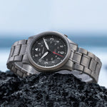 This Berny watch is elegantly displayed on a rocky surface with a background of the sea. It features a titanium bracelet and a black dial with both 12-hour and 24-hour markers. The watch combines durability and style, making it suitable for adventurous settings.